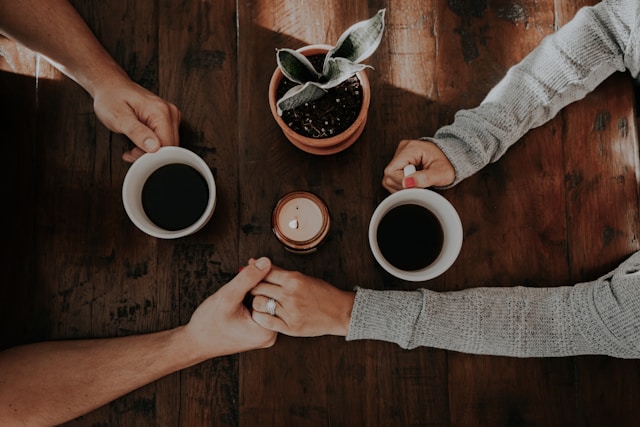 Two people sitting across from each other holding hands and drinking coffee